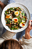 Top view of crop unrecognizable female in sweater eating delicious healthy dish with chickpeas boiled eggs and herbs during lunch in kitchen