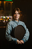 Glad female waitress with round tray looking at camera while standing near bar with assorted alcohol drinks in modern cafe