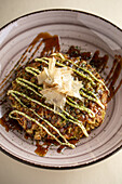 From above of tasty traditional Japanese okonomiyaki dish with sauce topped with bonito flakes served in ceramic bowl on light background