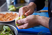 Unrecognizable male cook in uniform peeling fresh artichoke near bowl over table while working in restaurant