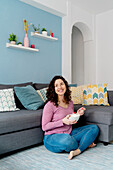 Smiling female with bowl eating breakfast while sitting on sofa in light room looking away