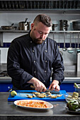 Bearded male chef cutting fresh artichokes while preparing delectable dish on table in kitchen of contemporary restaurant