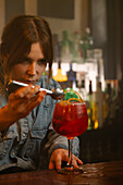 Focused female barkeeper adding lemon zest into wineglass with alcoholic red old fashioned cocktail while working at wooden bar counter