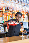 Focused African American female barkeeper pouring alcohol in glass with ice cube while making cocktail in bar