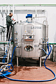 Side view of male engineer examining stainless steel tank with hoses in brewery with wet floor