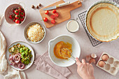 Top view of unrecognizable woman using whisk to beat raw chicken eggs in bowl near fresh ingredients and crust while preparing quiche in kitchen at home