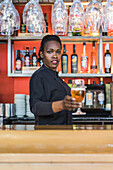 Cheerful black female barkeeper serving glass of refreshing beer in bar looking at camera