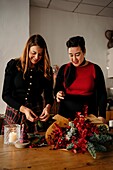 Fröhliche Freundinnen stehen am Tisch mit Kerzen und basteln kreative Weihnachtssträuße für die Weihnachtsfeier