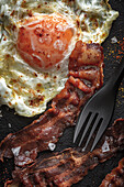 From above of sunny side up egg with fried bacon slices and condiments on tray against cutlery on dark background