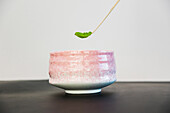 Wooden chashaku of dried green tea adding to ceramic bowl placed on table against white background