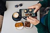 High angle of crop anonymous female blogger recording video of late coffee and cakes on cellphone in outdoor cafe