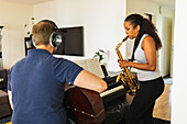 Hispanic Frau mit langen lockigen Haaren stehen und spielen Saxophon in der Nähe von positiven männlichen in Kopfhörer sitzen und spielen akustische Gitarre in hellen Raum in der Tageszeit