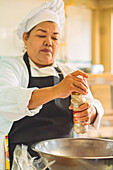 Focused female chef in uniform and apron adding spices into metal bowl while preparing food in kitchen of restaurant