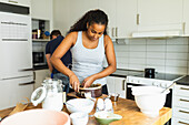 Young African American female standing at wooden table with ingredients and various cooking utensils placed on while covering cake mold with baking paper with unrecognizable man on background