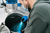From above side view of male engineer with blue light checking up metal vessel in brewery during pandemic
