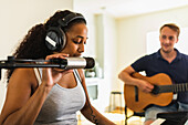 Ruhige hispanische Frau mit Kopfhörern, die E-Piano spielt und in ein Mikrofon singt, während sie mit einem Freund, der Akustikgitarre spielt, tagsüber zu Hause Musik macht
