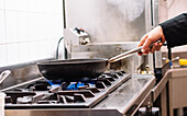 Side view of crop unrecognizable cook preparing dish in frying pan on stove in kitchen of restaurant