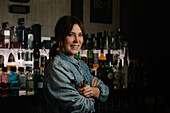 Attractive smiling female bartender with jigger and tong in hands looking at camera while standing in bar with various alcohol drinks