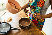 From above crop hands of unrecognizable female in apron pouring liquid chocolate batter into baking form with baking paper