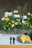 Blick von oben auf einen Teller und eine Schüssel mit Besteck und Serviette, die auf einem mit zarten frischen weißen und gelben Blumen dekorierten Tisch stehen