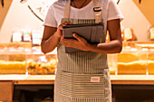 Cropped unrecognizable black female vendor in uniform with tablet smiling and inspecting pasta in glass container during work in zero waste grocery store looking away