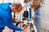 Side view of male worker in mask with cellphone checking up liquid in tube of lauter tun in beer factory during covid 19 pandemic