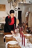 Calm young woman in casual clothes standing and putting burning candle on festive table in light room in daytime
