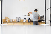 Back view of anonymous male barista pouring coffee beans from bag into modern professional coffee grinder at wooden counter in coffee house