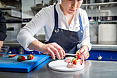 Woman in apron using tweezers to put slice of fresh strawberry on top of whipped cream while cooking dessert in kitchen of contemporary restaurant