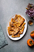 Top view of white plate with delicious French toasts placed near jar with honey and fresh persimmons on gray table