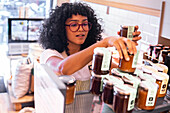 From above young African American female vendor in glasses arranging glass jars with various jam sauces on shelf during work in eco friendly grocery shop