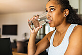 Young positive black female with curly hair drinking fresh pure water from glass and looking away at home