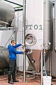 Side view of male worker in sterile mask pouring fermented beer from metal vessel into glass in factory