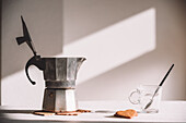 Metal moka pot with opened lid placed on white table near glass cup and heart shaped cookies on sunny day