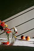 Glasvasen mit blühenden Tulpen und Nelken auf einem Tisch in der Nähe von Äpfeln mit Schatten in einer Wand