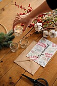 From above of crop unrecognizable person putting burning candle on wooden table with postcard and decorative bouquet