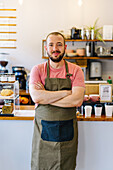 Positive male barista in apron looking at camera with crossed arms while standing near counter with paper cups and desserts in coffee house
