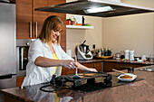 Side view of cheerful adult woman in white shirt standing at counter and flipping pancakes with spatula while making breakfast
