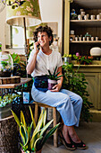 Smiling female florist with potted plant sitting in flower shop while speaking on mobile phone and getting order