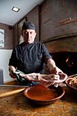 Male chef with raw poultry standing near table with bowl during work in kitchen of restaurant with furnace