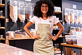 Self assured young African American woman in uniform and glasses with curly hair with hands on waist looking at camera during work in zero waste grocery shop