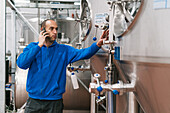 Side view of male entrepreneur talking on cellphone against stainless steel vessels on wet floor in beer factory