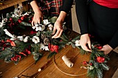 Unkenntlich gemachte Freundinnen stehen an einem Holztisch und basteln einen kreativen Weihnachtskranz aus dekorativen Tannen und roten Bändern für die Weihnachtsfeier