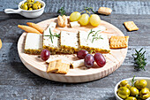 Appetizing cheese served on wooden table with ripe grapes and crackers decorated by rosemary sprigs near olives in bowls on table