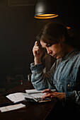 Side view of focused female worker using calculator to count income while sitting at counter with documents during work in cafe