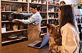 Side view of calm woman seller in apron standing and demonstrating glass jar with nuts to female client standing with dog in hand at wooden counter in eco friendly shop