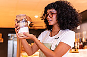 Happy young ethnic woman in uniform and glasses holding glass jar with nuts and diverse flours while working in zero waste grocery shop