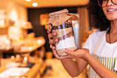 Cropped unrecognizable happy young ethnic woman in uniform and glasses holding glass jar with nuts and diverse flours while working in zero waste grocery shop