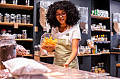 Glad young African American woman in striped apron and glasses smile and putting glass box of dried mango on shelf during work in zero waste grocery store