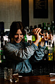 Cheerful female bartender making tasty alcohol cocktail with shaker while standing at counter with glasses in bar with various beverages